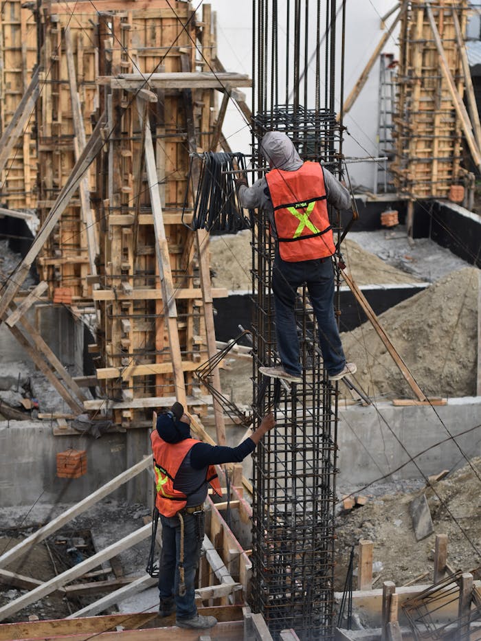 services-img Two workers in safety vests climb a steel structure on a busy construction site.