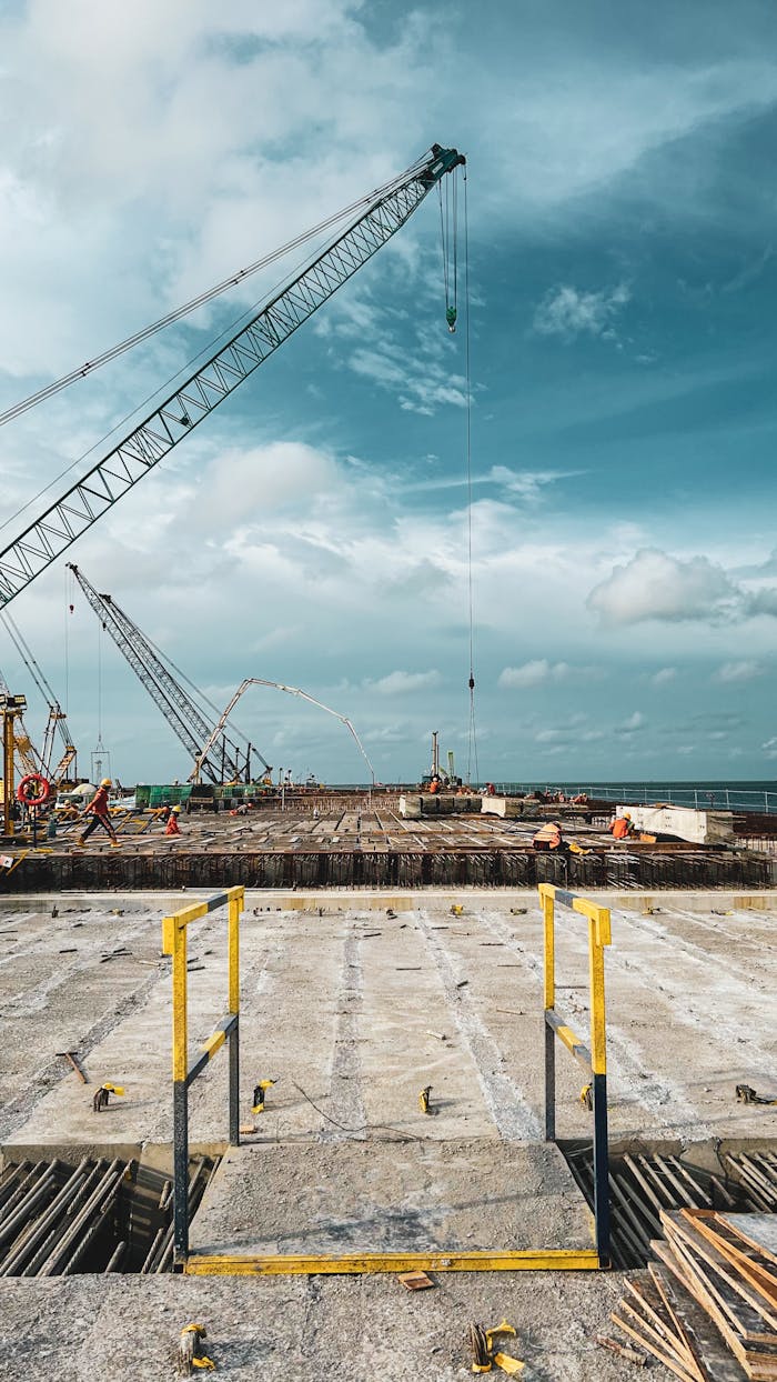 about-img A bustling construction site by the sea with cranes and workers under a vibrant blue sky.